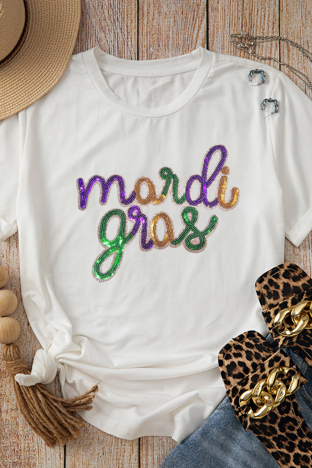 White t-shirt with 'mardi gras' in colorful letters on a wooden surface with accessories.