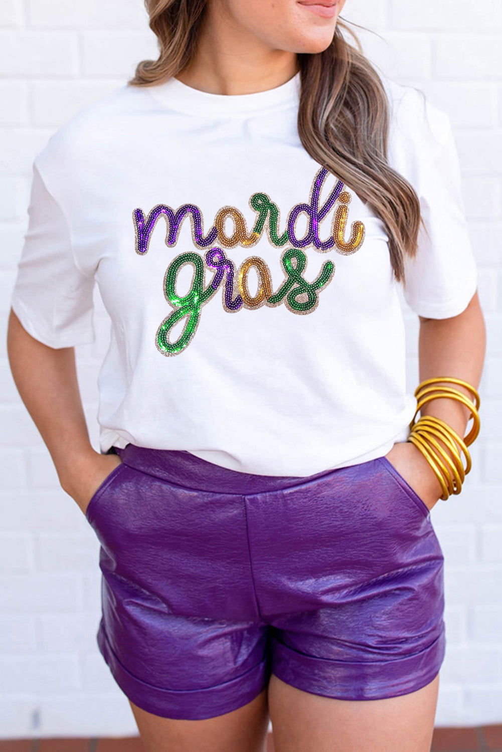 Person wearing a white t-shirt with 'mardi gras' text and purple shorts against a white brick wall.