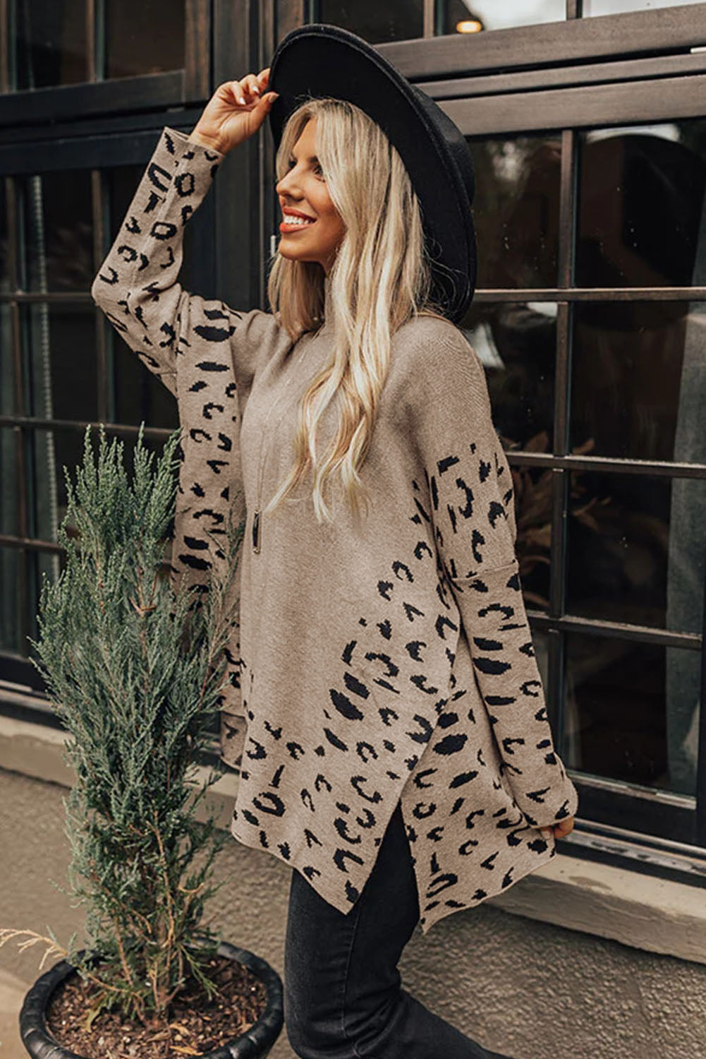 Woman wearing a leopard print cardigan and black hat standing next to a plant.