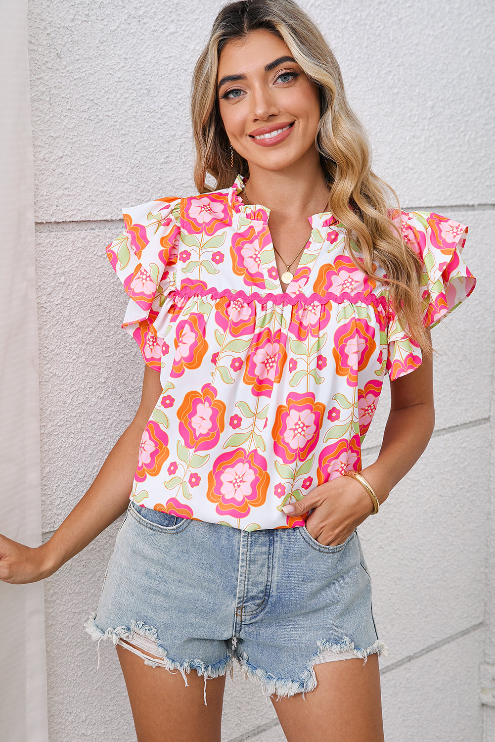 Woman wearing a colorful floral blouse and denim shorts against a light gray wall.