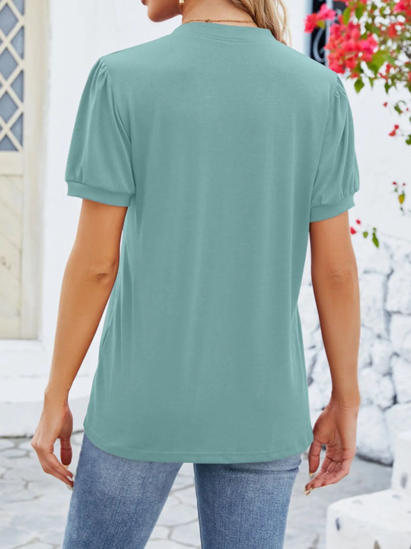 Person wearing a light green t-shirt and blue jeans standing outdoors with a white wall and floral decorations in the background.