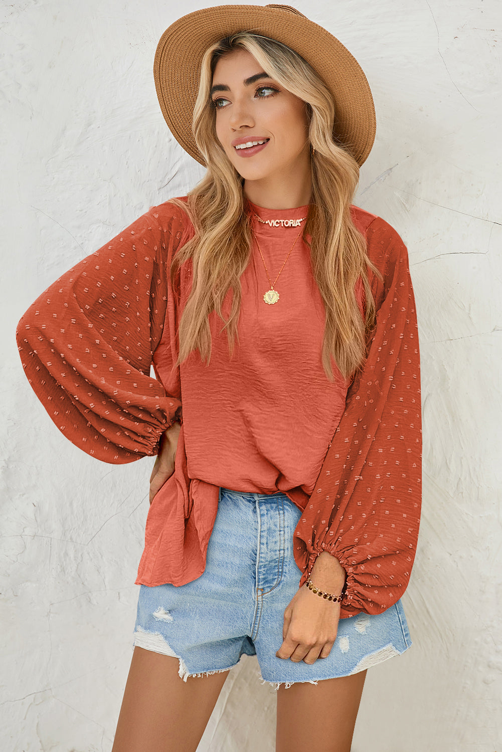 Woman wearing a rust-colored blouse with long sleeves and denim shorts against a white wall.