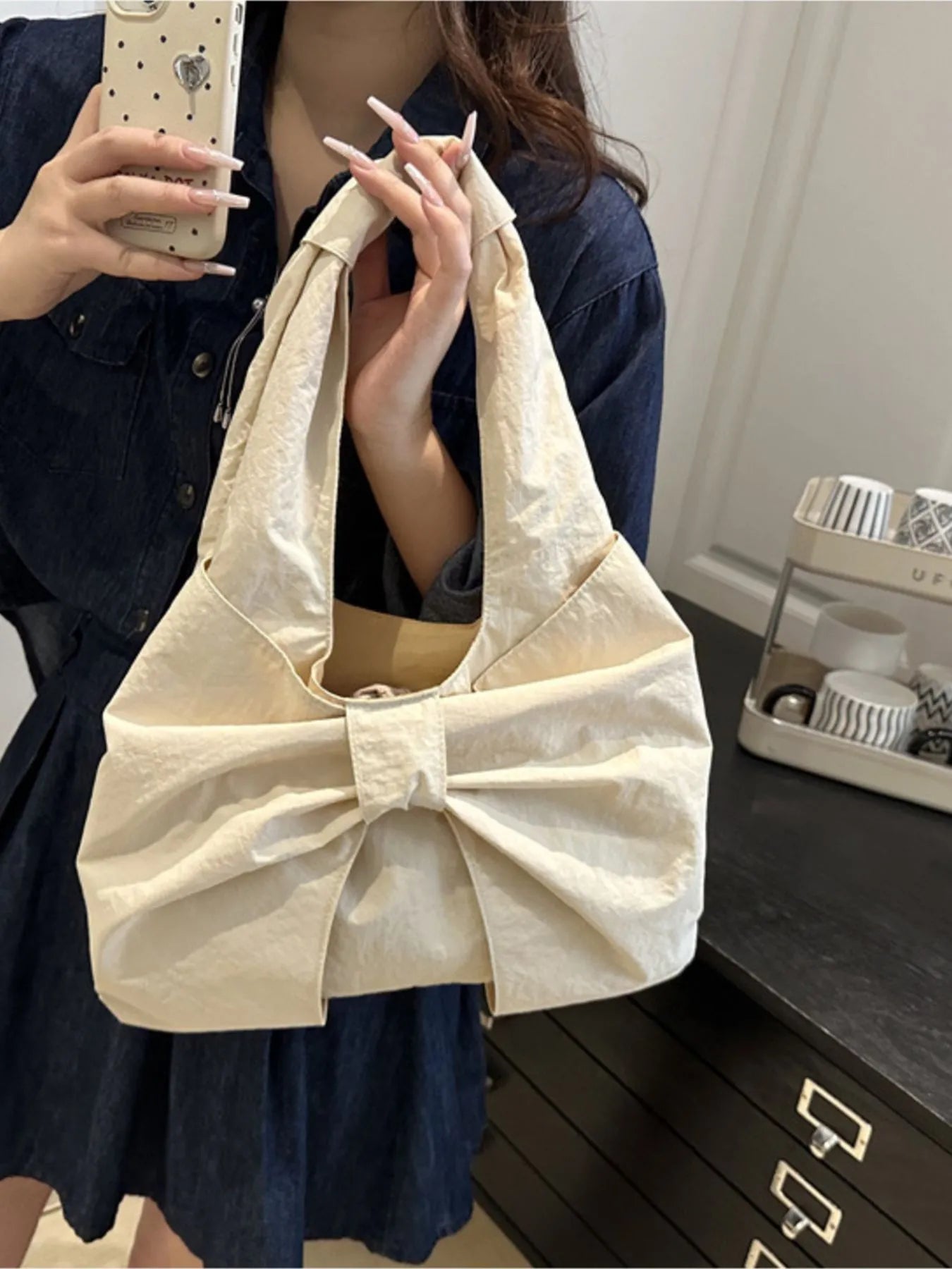 Person holding a beige handbag with a large bow, taking a mirror selfie.