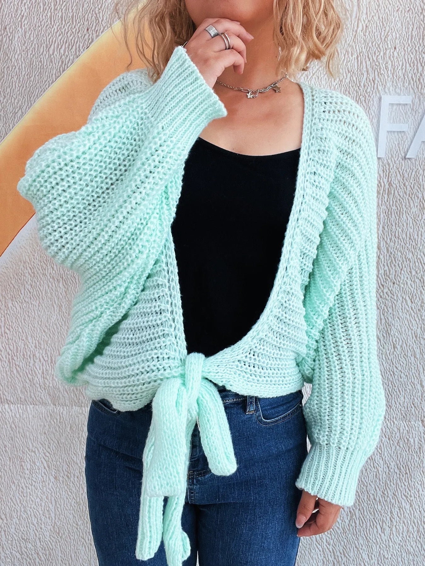 Person wearing a light blue knitted cardigan over a black top and blue jeans against a plain background