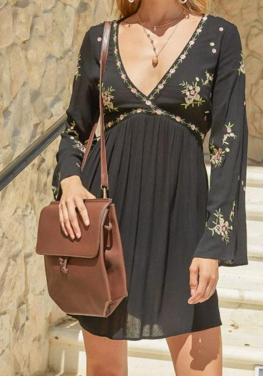 Woman wearing a black floral dress holding a brown leather bag against a stone wall.
