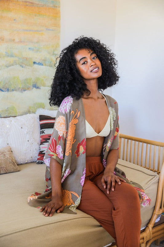 Woman sitting on a couch wearing a floral kimono and brown pants, with a colorful abstract painting in the background.