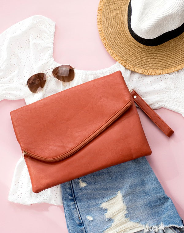 Orange clutch bag with sunglasses, white lace top, straw hat, and denim jeans on a pink surface.