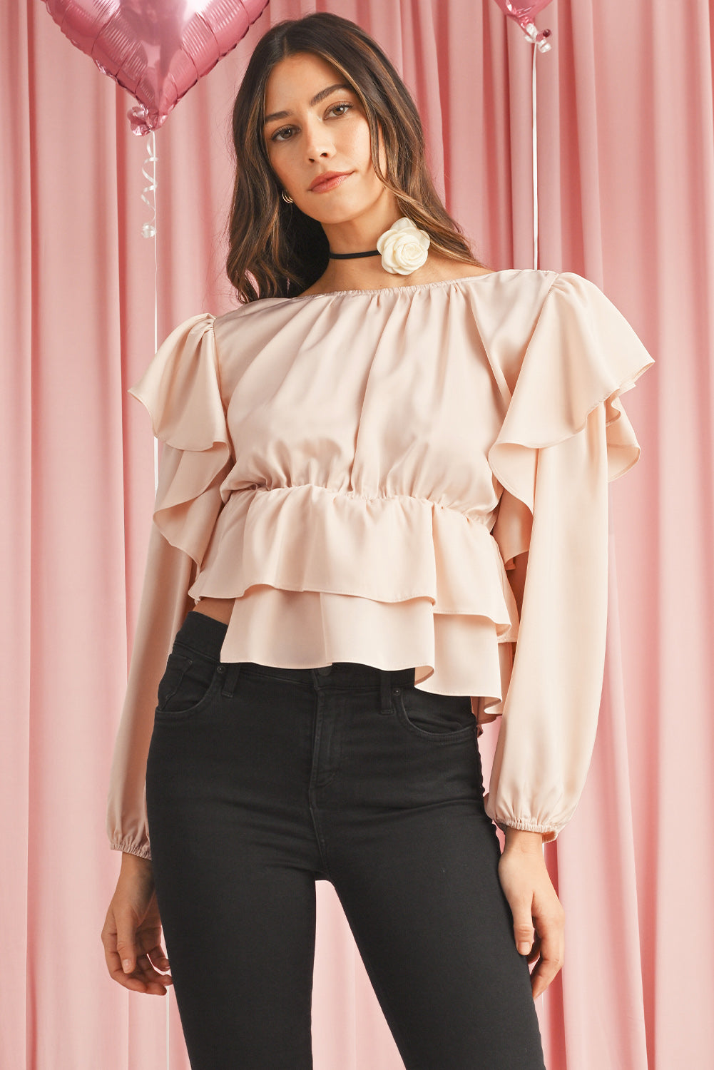 Woman wearing a light pink off-shoulder top with ruffled layers against a pink curtain background.