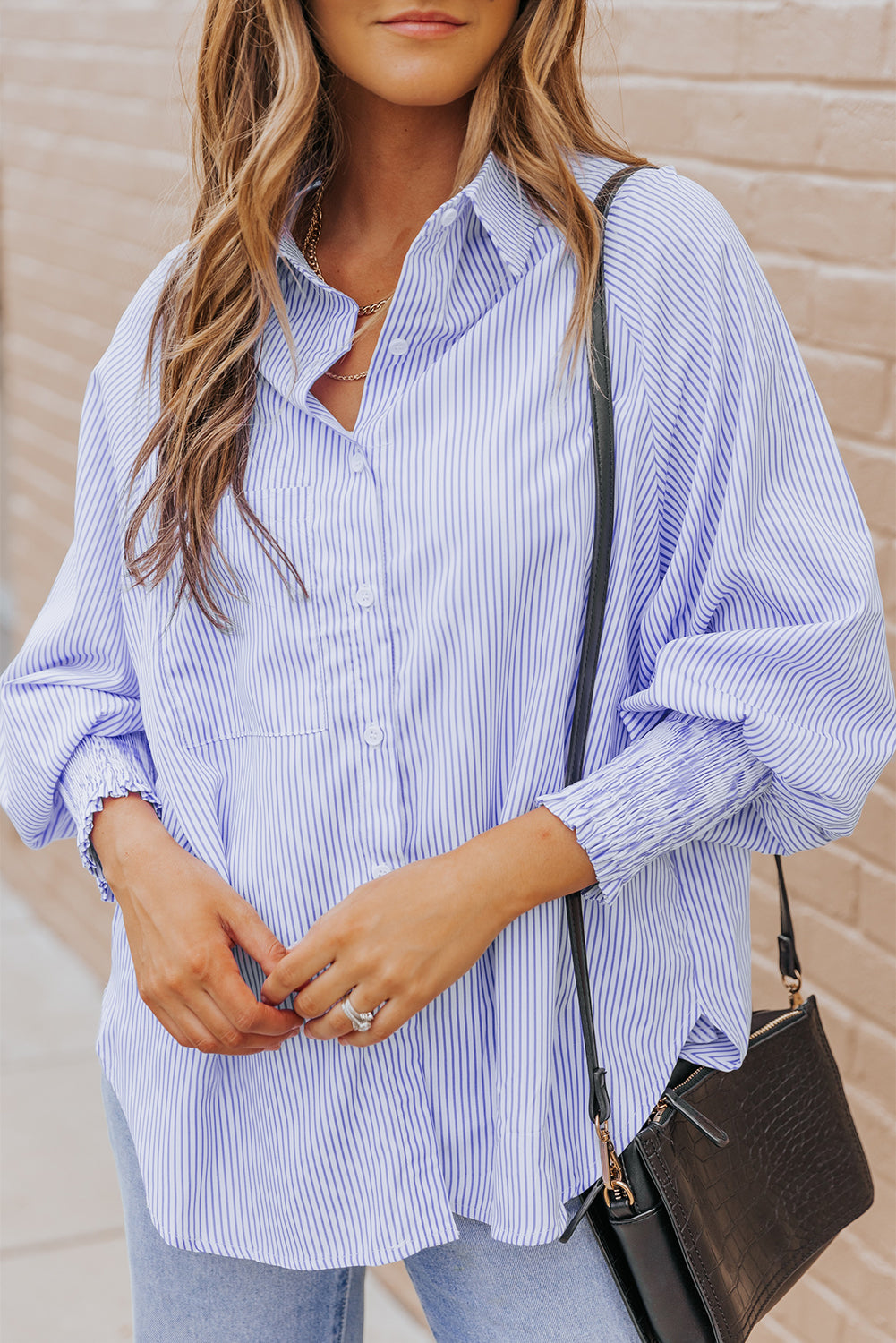 Woman wearing a light blue striped shirt with rolled-up sleeves, holding a black handbag against a beige wall.