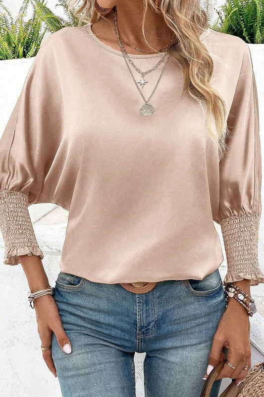 Woman wearing a beige blouse with smocked cuffs and blue jeans outdoors.