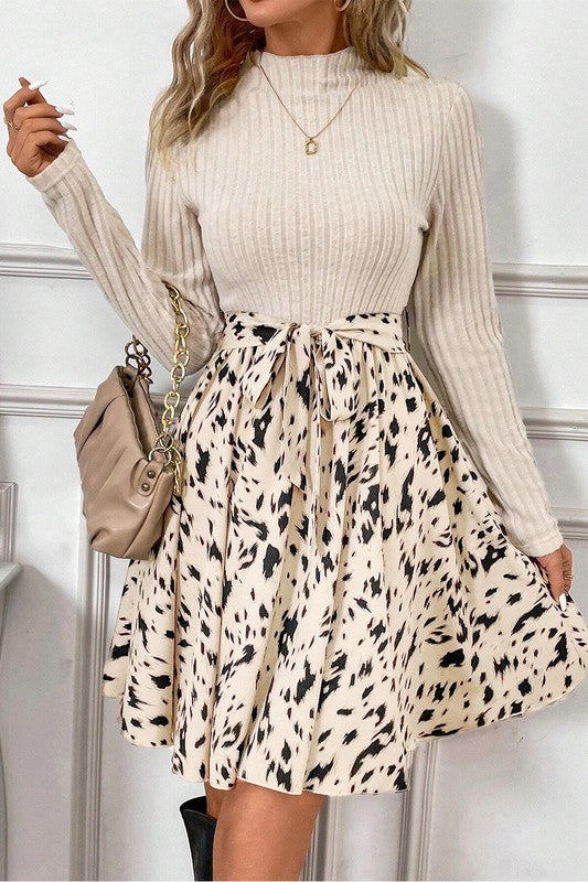 Woman wearing a beige ribbed sweater and leopard print skirt with a beige handbag against a white wall.