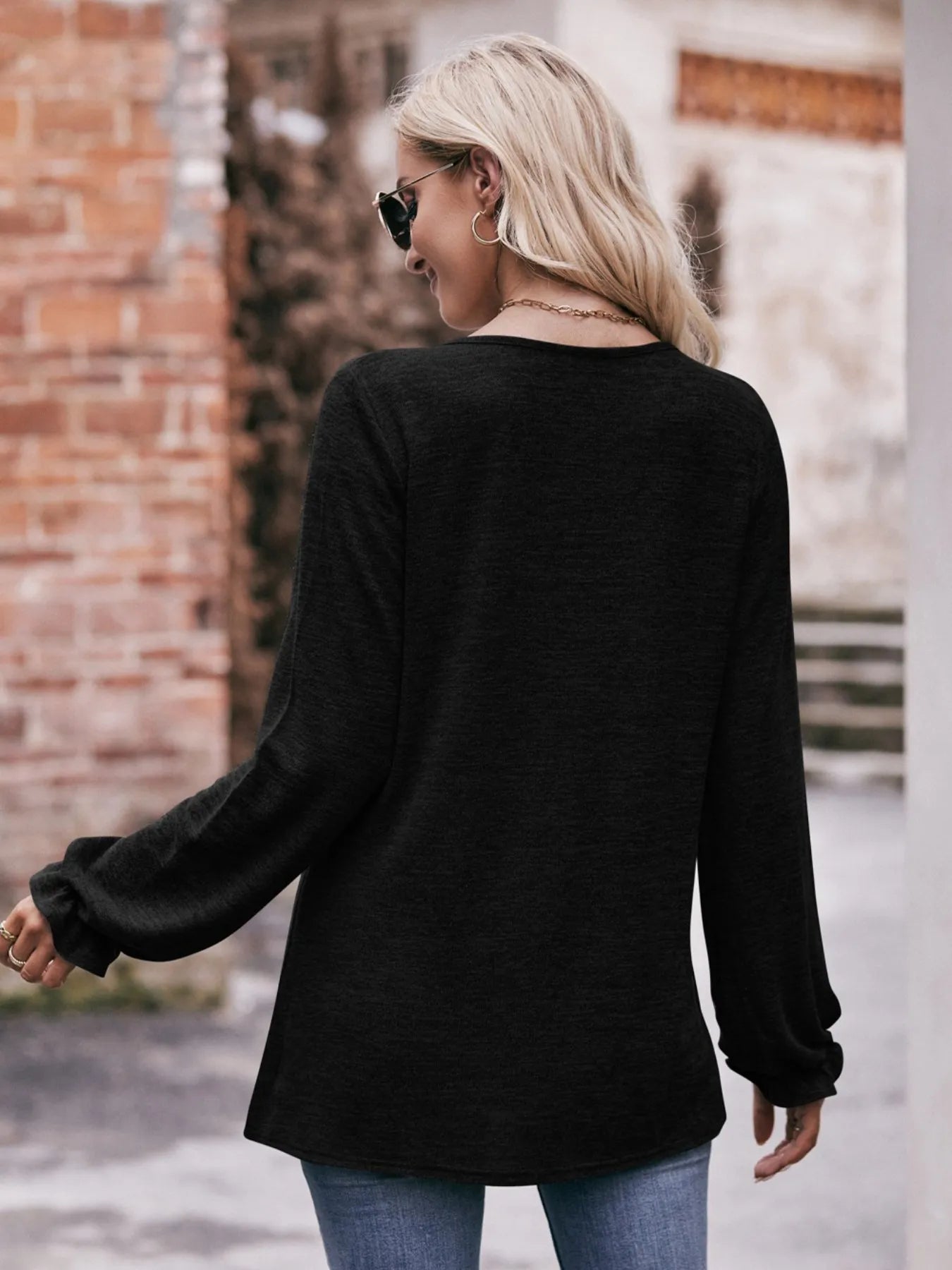 Woman wearing a black long-sleeve top and blue jeans, standing in front of a brick wall.