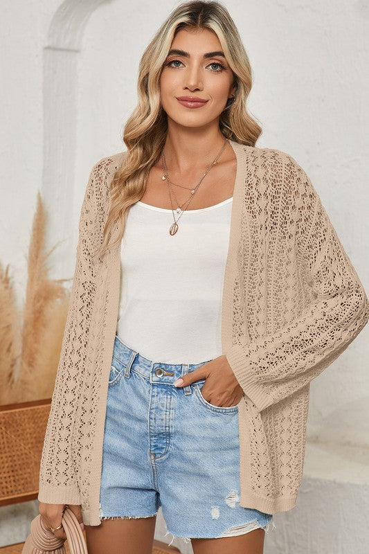 Woman wearing a beige lace cardigan over a white top and denim shorts, standing against a neutral background.