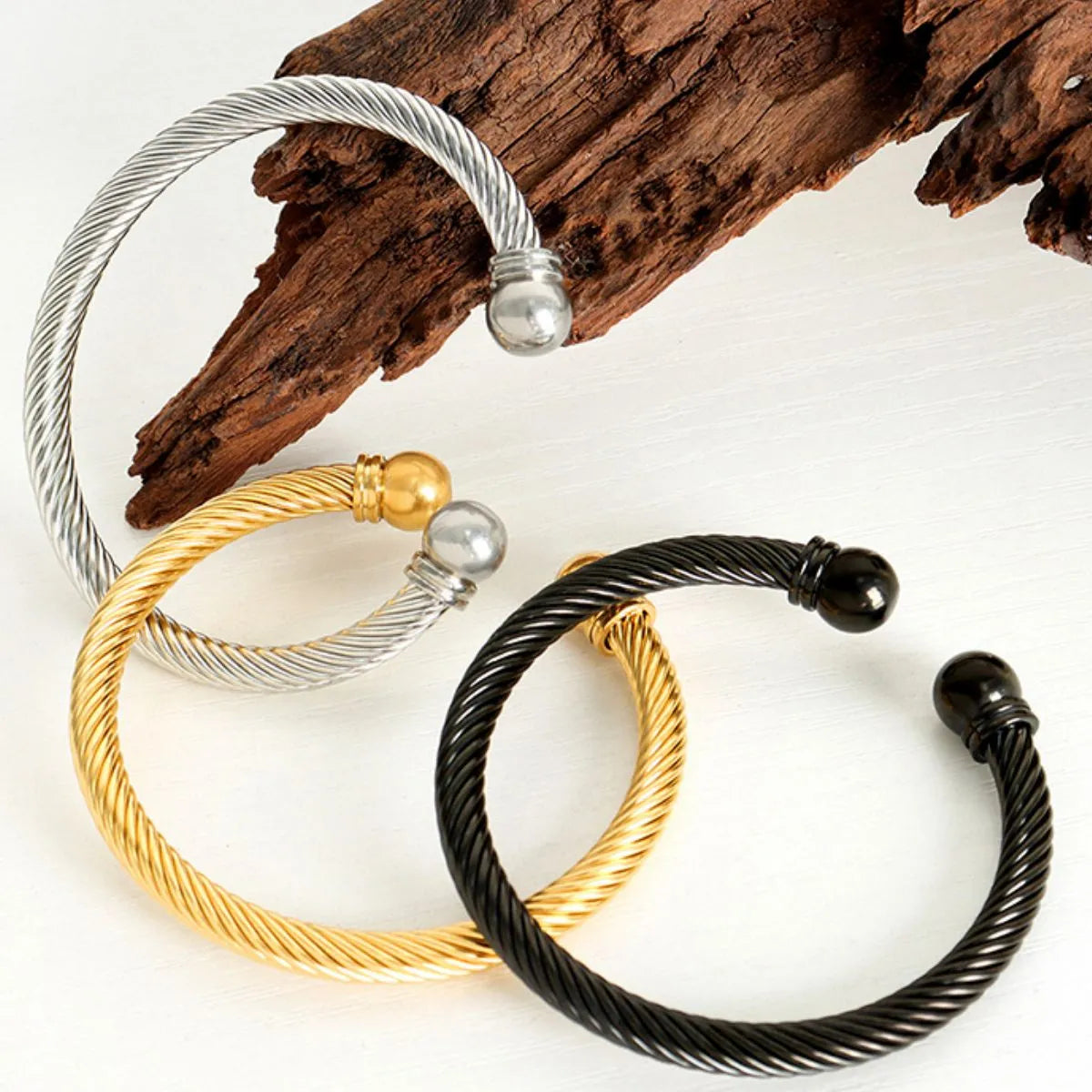 Three braided bracelets in silver, gold, and black on a white background with wooden log.