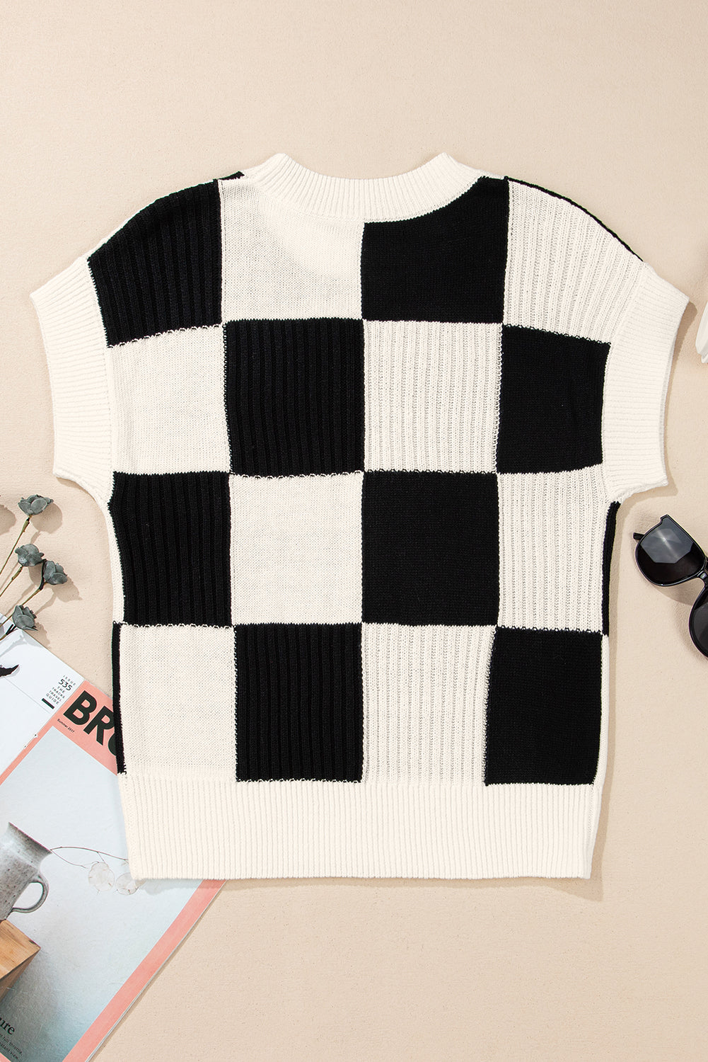 Black and white checkered sweater vest on a beige surface with magazines and sunglasses.