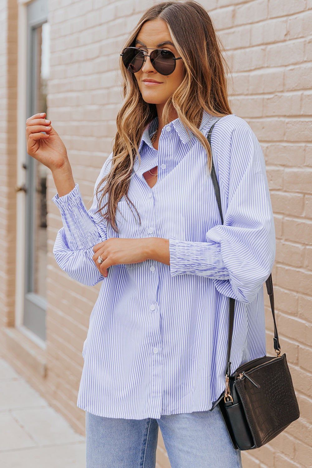 Woman wearing a light blue striped shirt and jeans, standing against a beige brick wall.