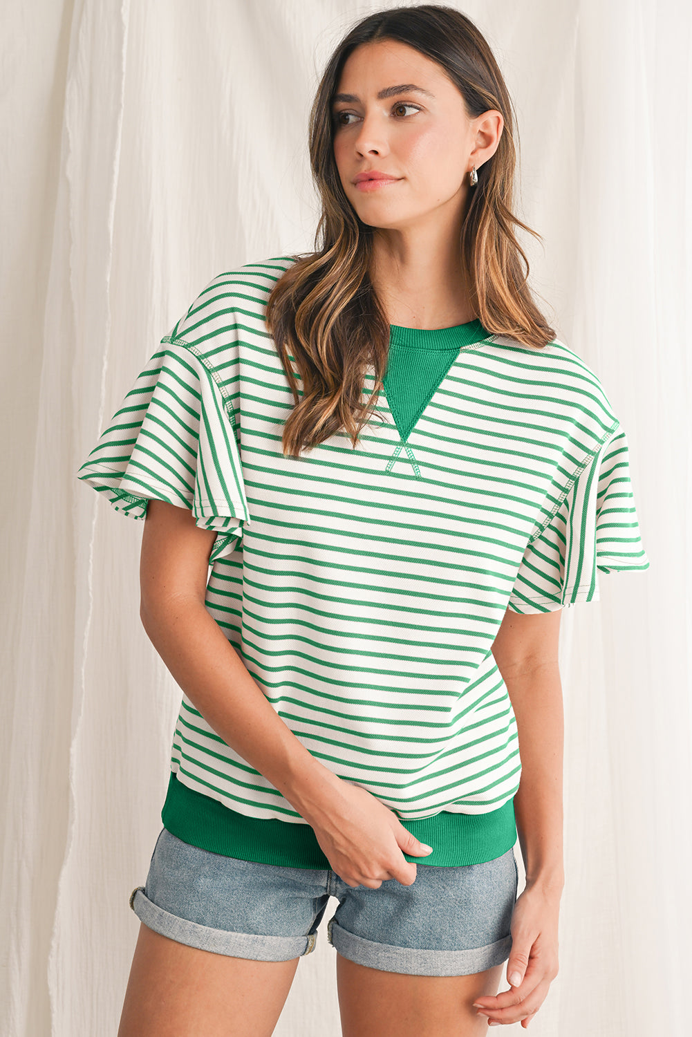 Woman wearing a green and white striped shirt with denim shorts against a white curtain background