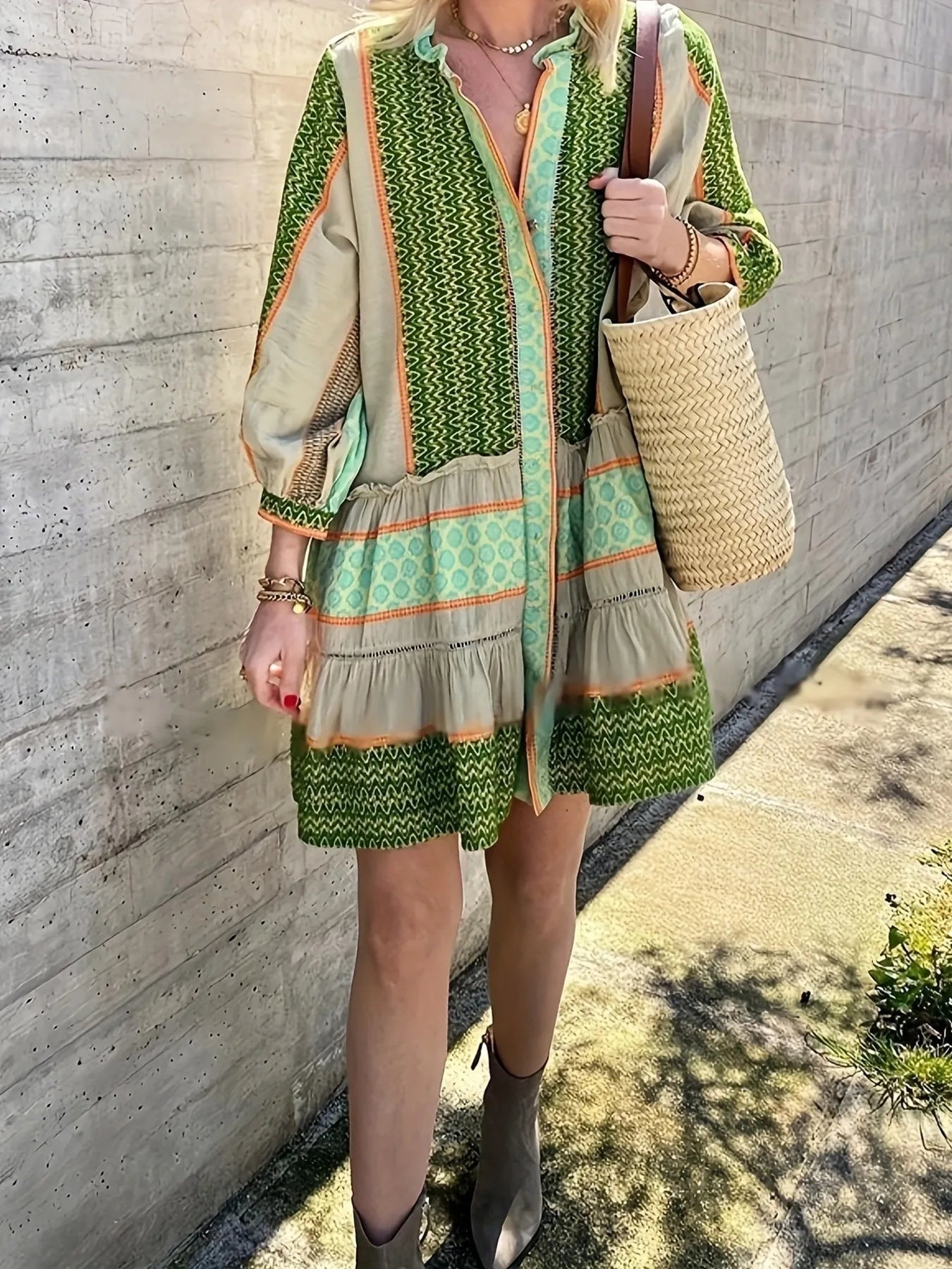 Person wearing a green and beige patterned dress holding a straw bag against a concrete wall.