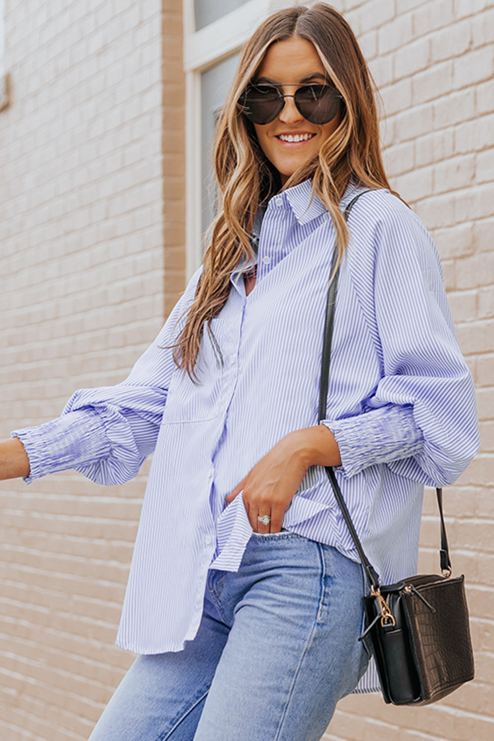 Woman wearing a light blue plaid shirt and jeans, standing against a beige brick wall.