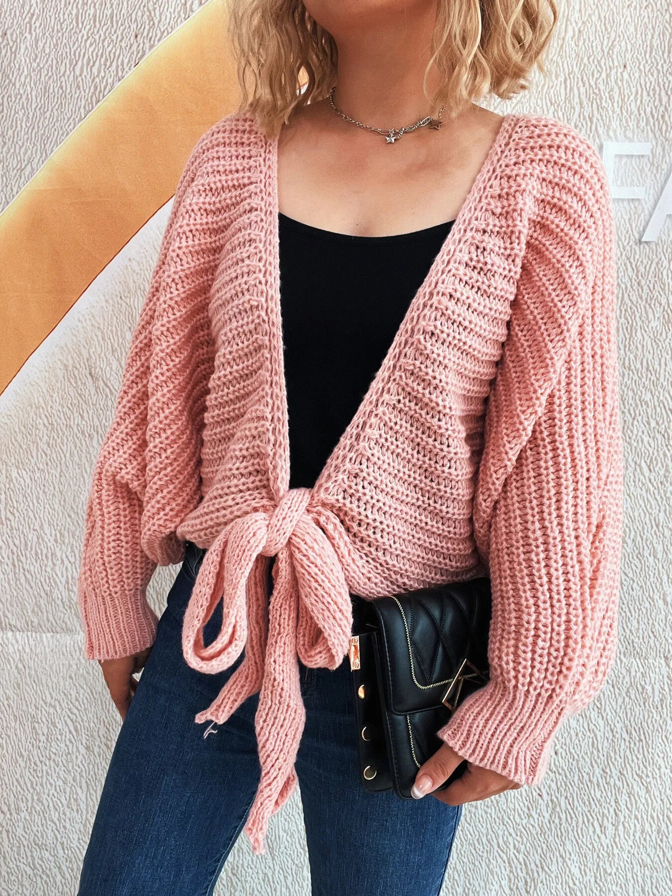 Person wearing a pink knitted cardigan with a tie front, black top, and blue jeans against a light background.