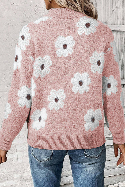 Pink sweater with white floral patterns worn by a person against a light gray wooden background