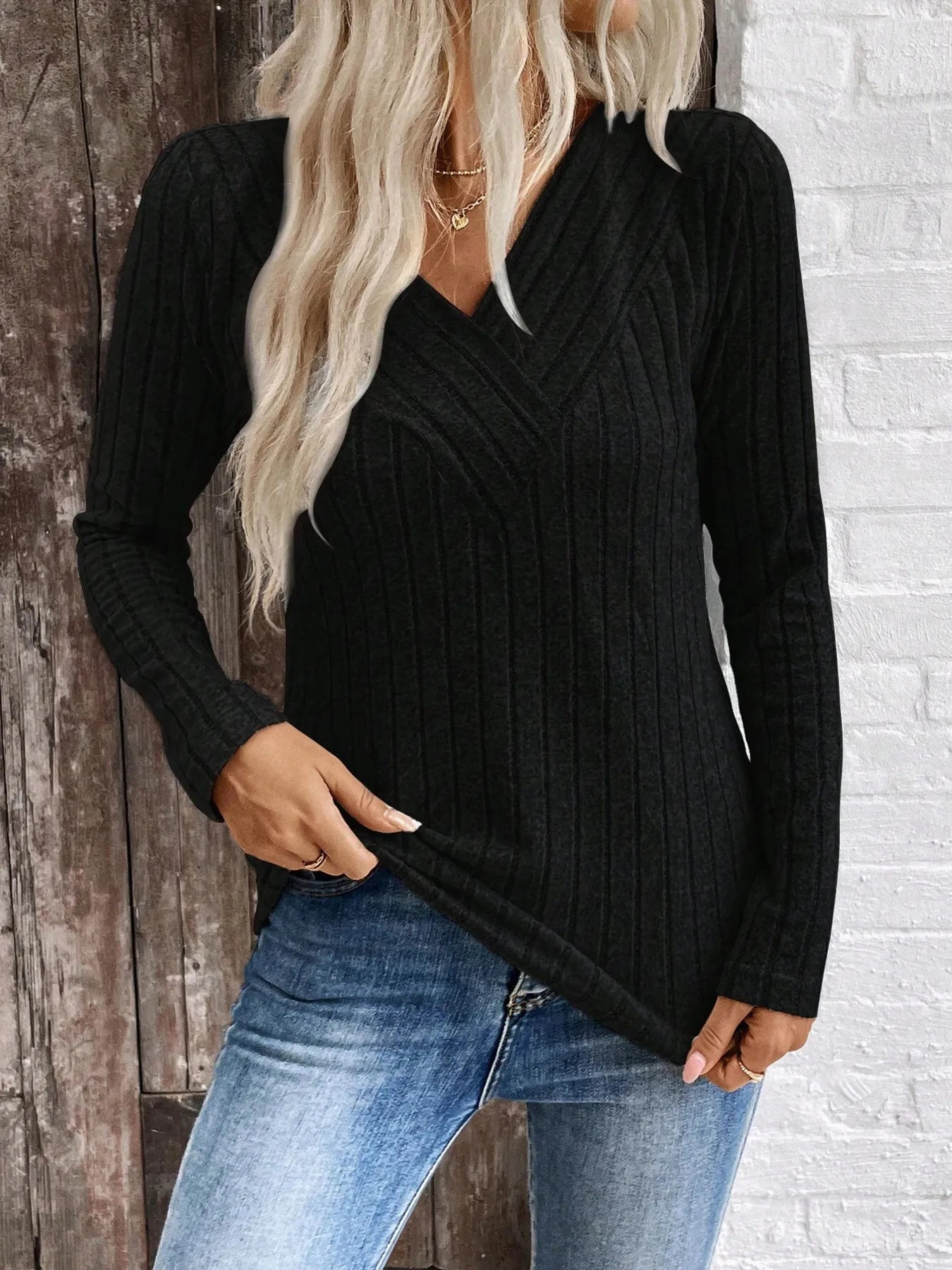Person wearing a black ribbed sweater and blue jeans against a wooden and white brick wall.