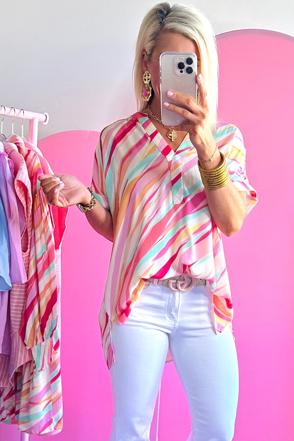 Person wearing a colorful striped shirt and white pants, standing in front of a pink background with a rack of clothes.
