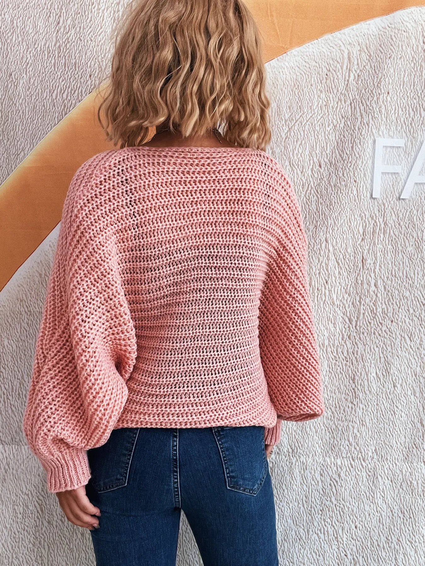 Person wearing a pink knitted sweater and blue jeans against a textured wall.