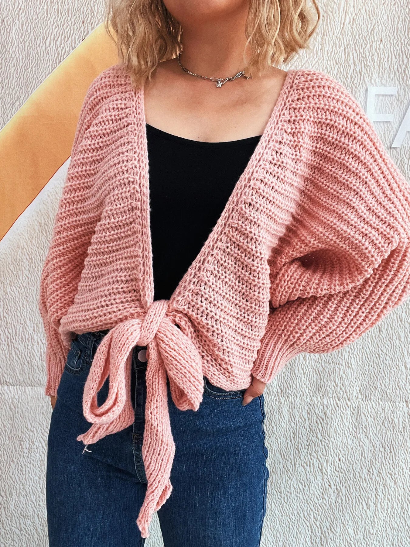 Person wearing a pink knitted cardigan over a black top and blue jeans against a light background