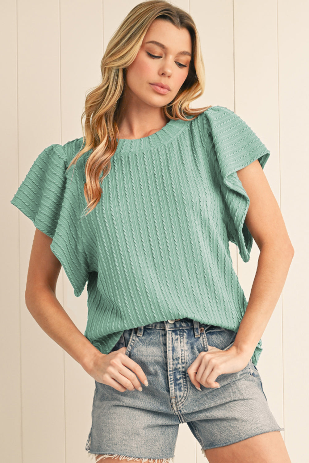 Woman wearing a green textured top and denim shorts against a neutral background