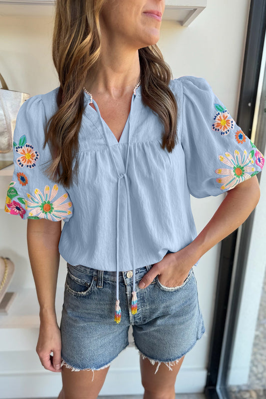 Woman wearing a light blue blouse with colorful floral embroidery and denim shorts indoors.