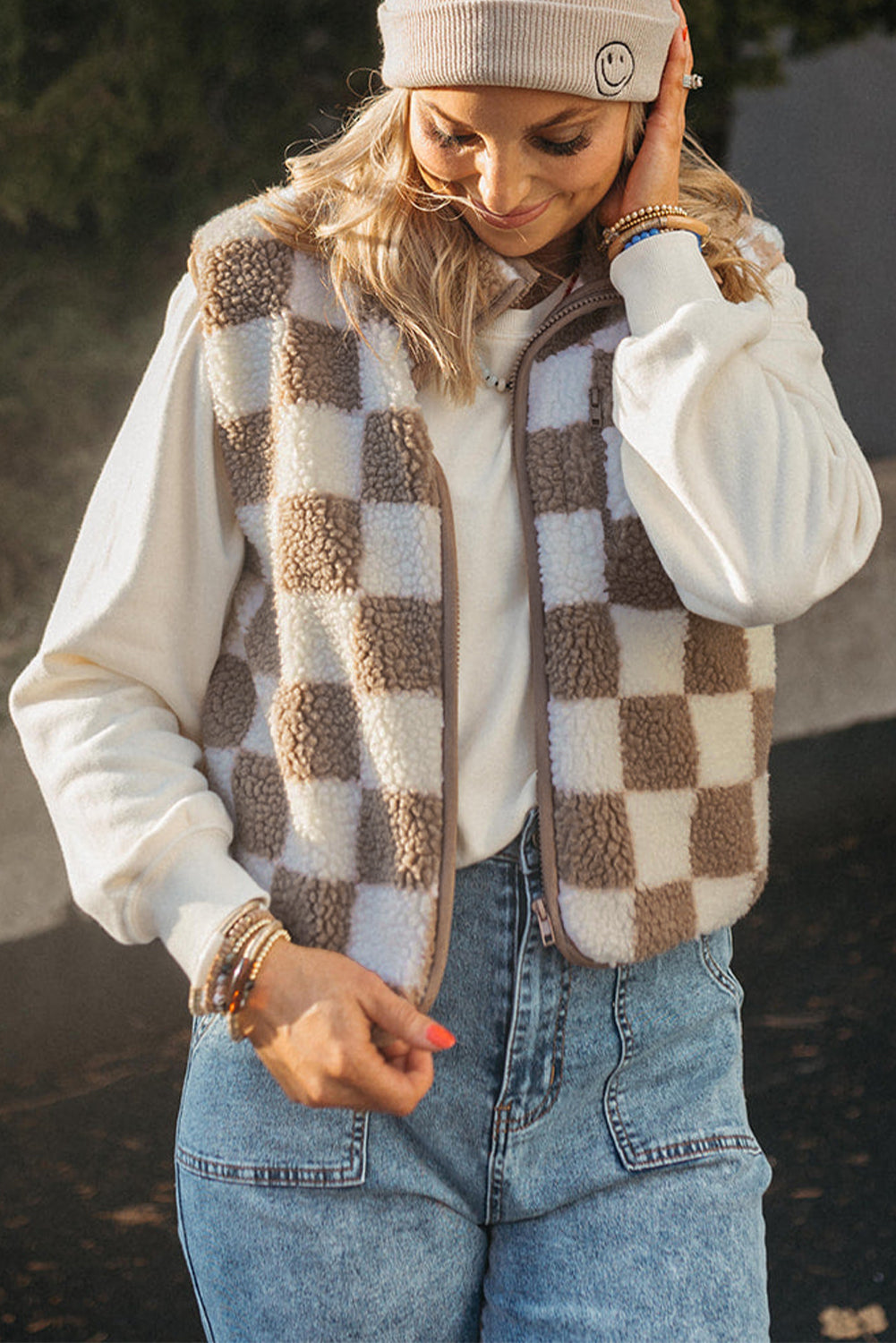 Person wearing a checkered vest, white sweater, and jeans outdoors.