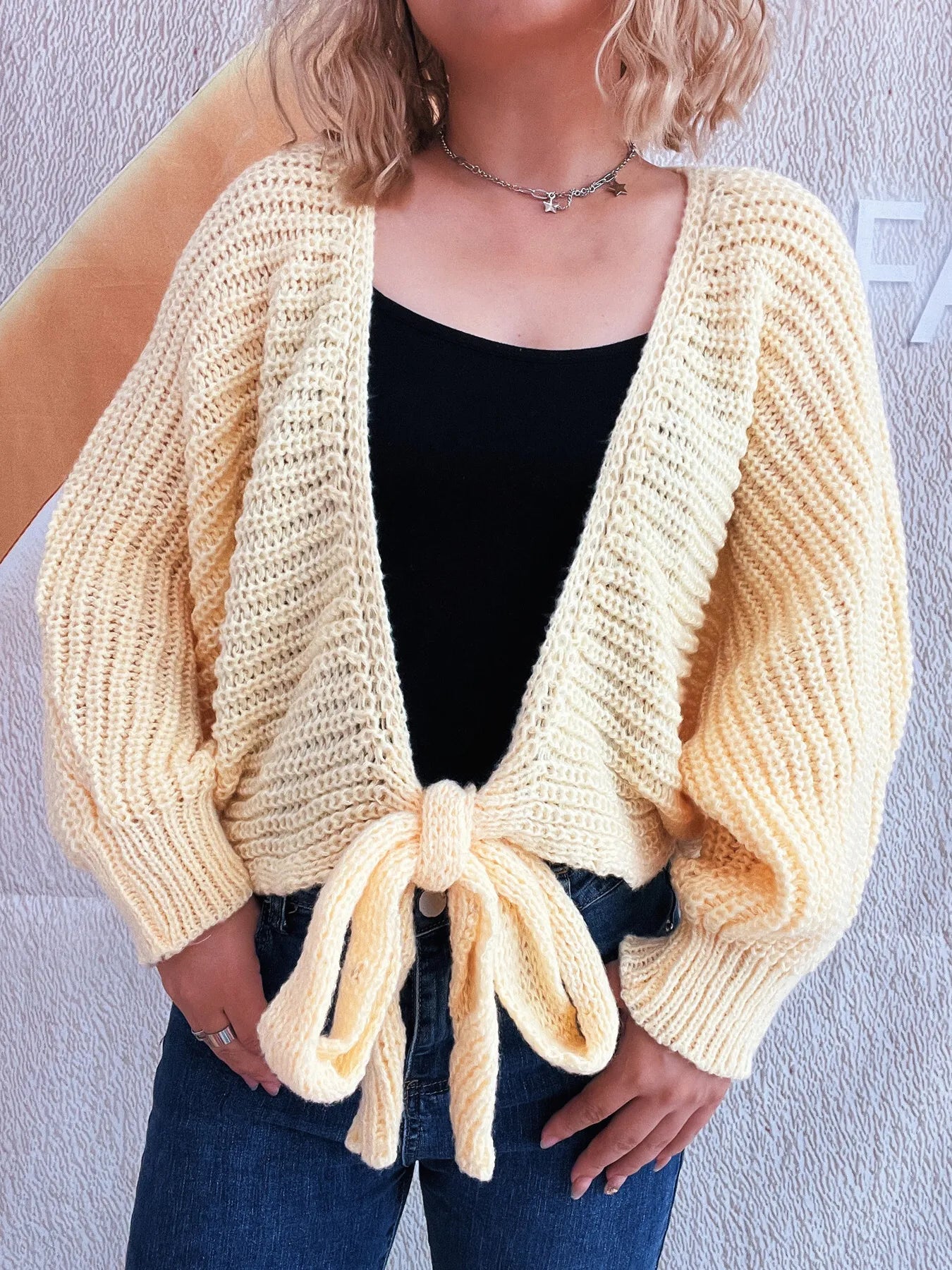 Person wearing a light yellow knitted cardigan over a black top and blue jeans against a plain background