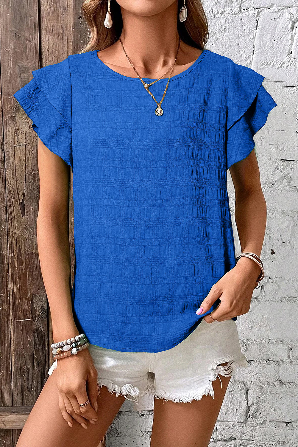 Woman wearing a blue top with ruffled sleeves against a wooden and white brick wall.