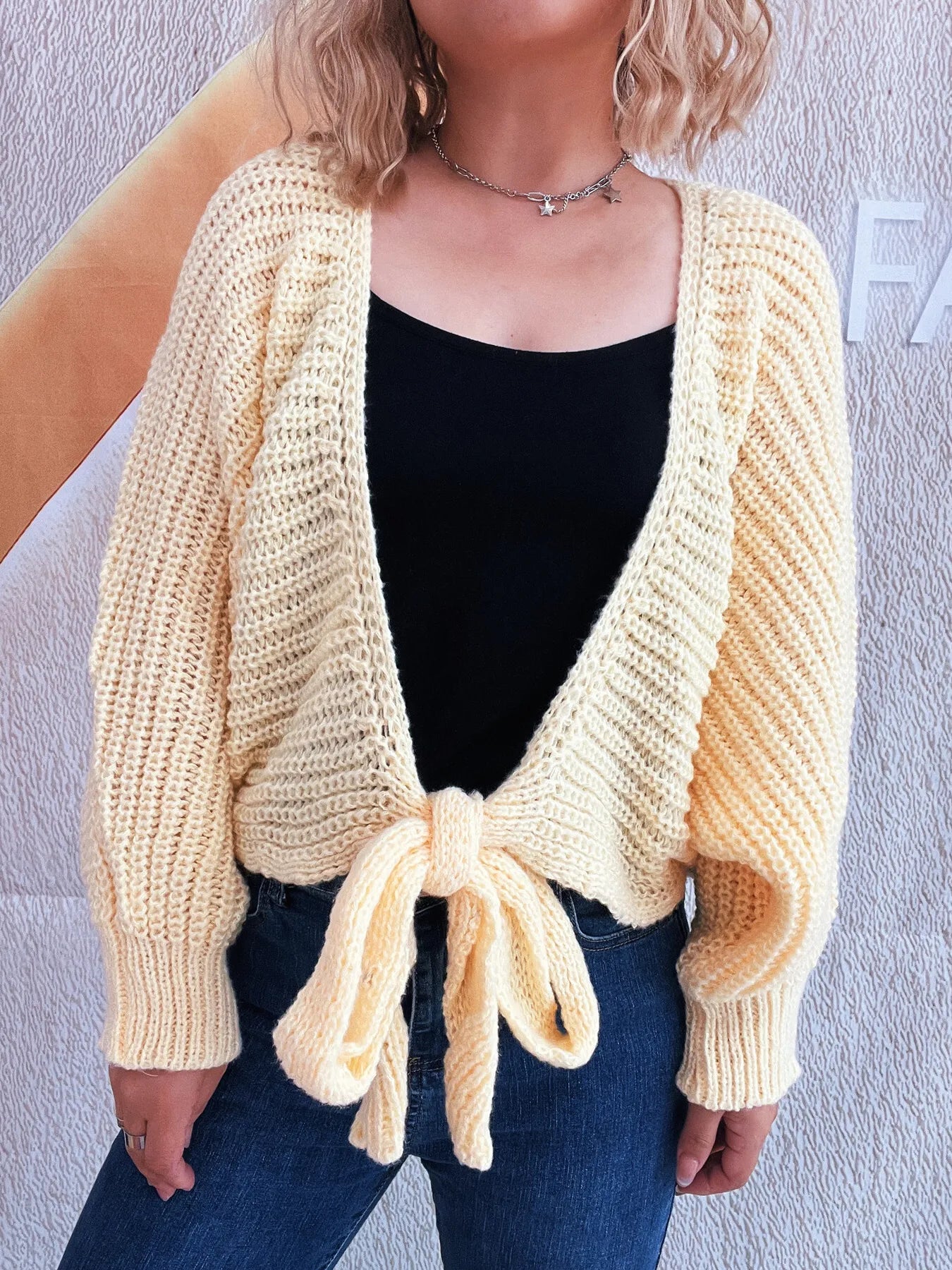 Person wearing a yellow knitted cardigan over a black top and blue jeans against a light gray background.