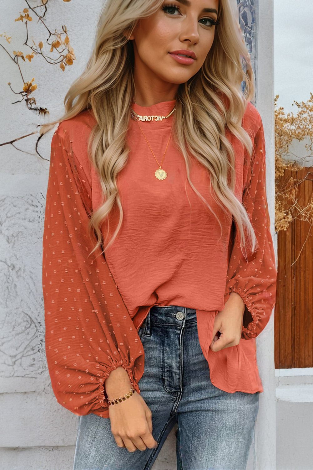 Woman wearing a rust-colored long-sleeve top with a subtle pattern and blue jeans, standing against a white wall with decorative elements.