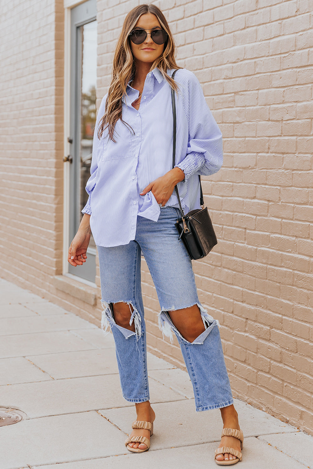 Woman wearing a light blue shirt and ripped jeans standing against a beige brick wall.