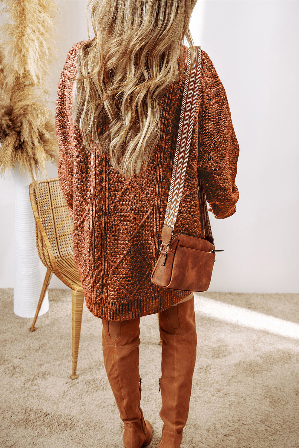 Person wearing a brown knitted sweater and matching over-the-knee boots, standing in a room with a chair and decorative items.