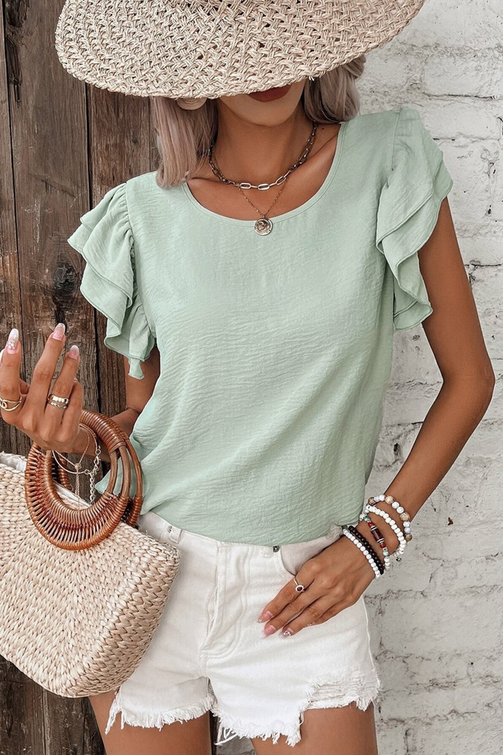 Woman wearing a light green ruffled shirt and white shorts, holding a straw bag against a wooden and white brick wall.