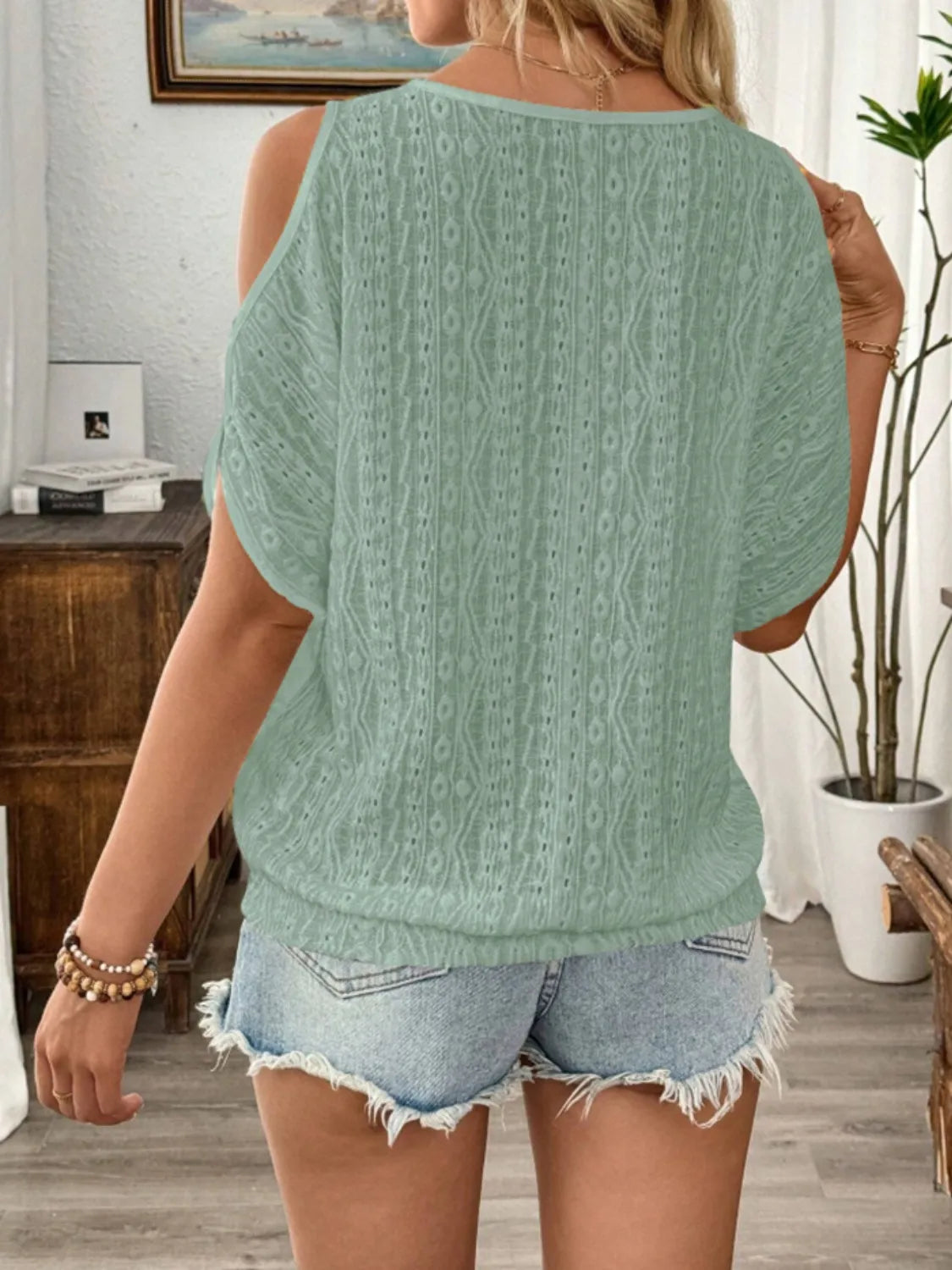 Person wearing a green cable knit sweater and denim shorts in a room with a desk and plant.