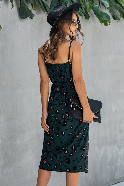 Woman wearing a dark green dress with red patterns and a black hat, standing against a gray wall with green leaves.