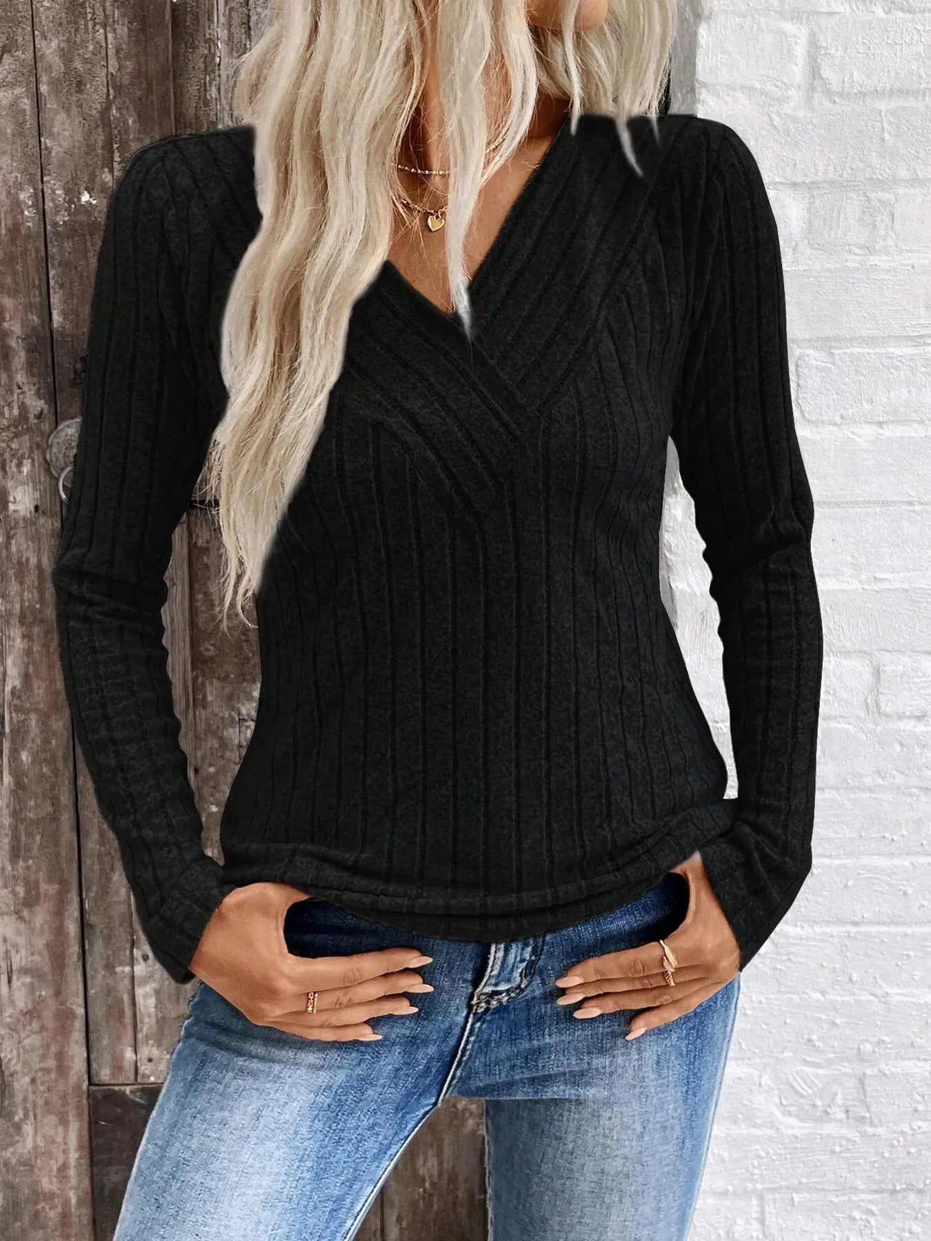 Person wearing a black ribbed sweater and blue jeans against a white brick wall.
