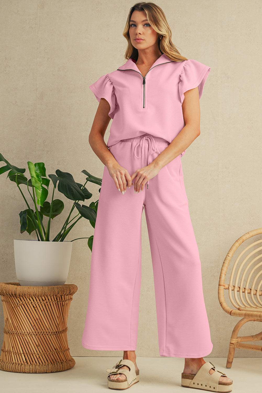 Woman wearing a pink outfit standing in a room with a plant and wicker chair.