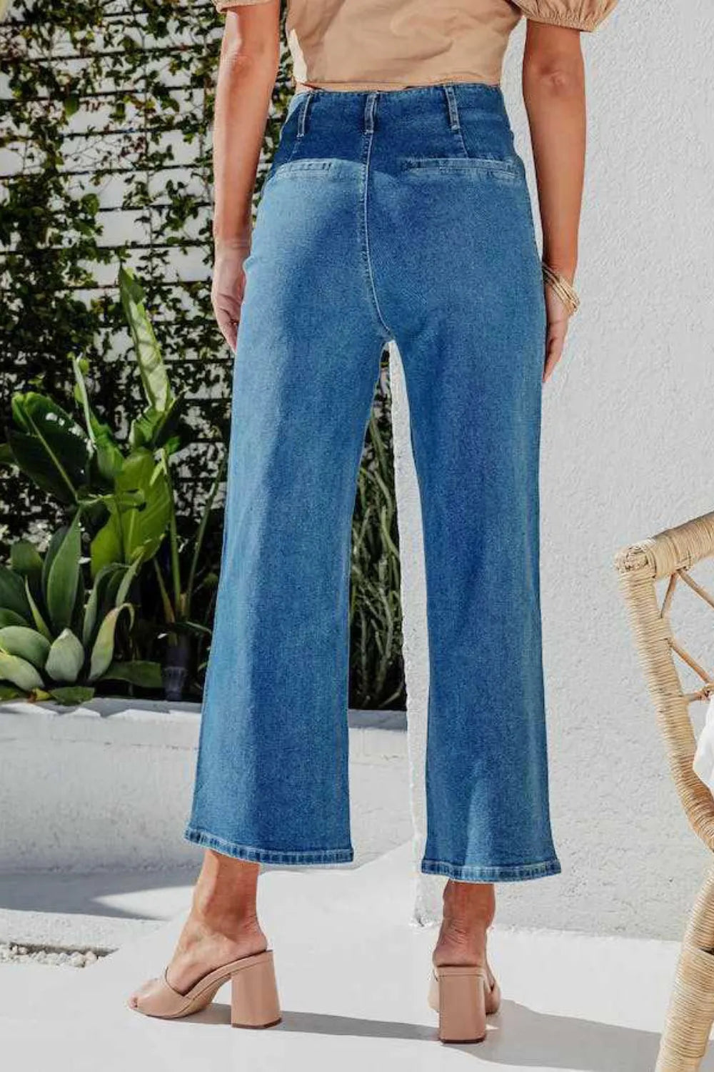 Person wearing blue jeans standing outdoors with plants and a chair in the background