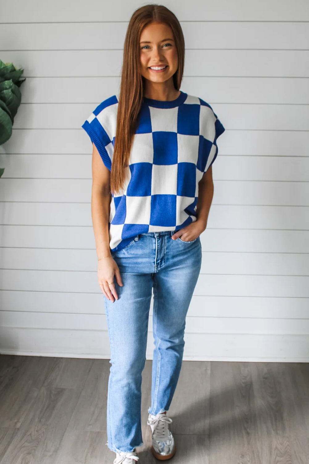 Woman wearing a blue and white checkered top and jeans standing against a white wooden panel background.