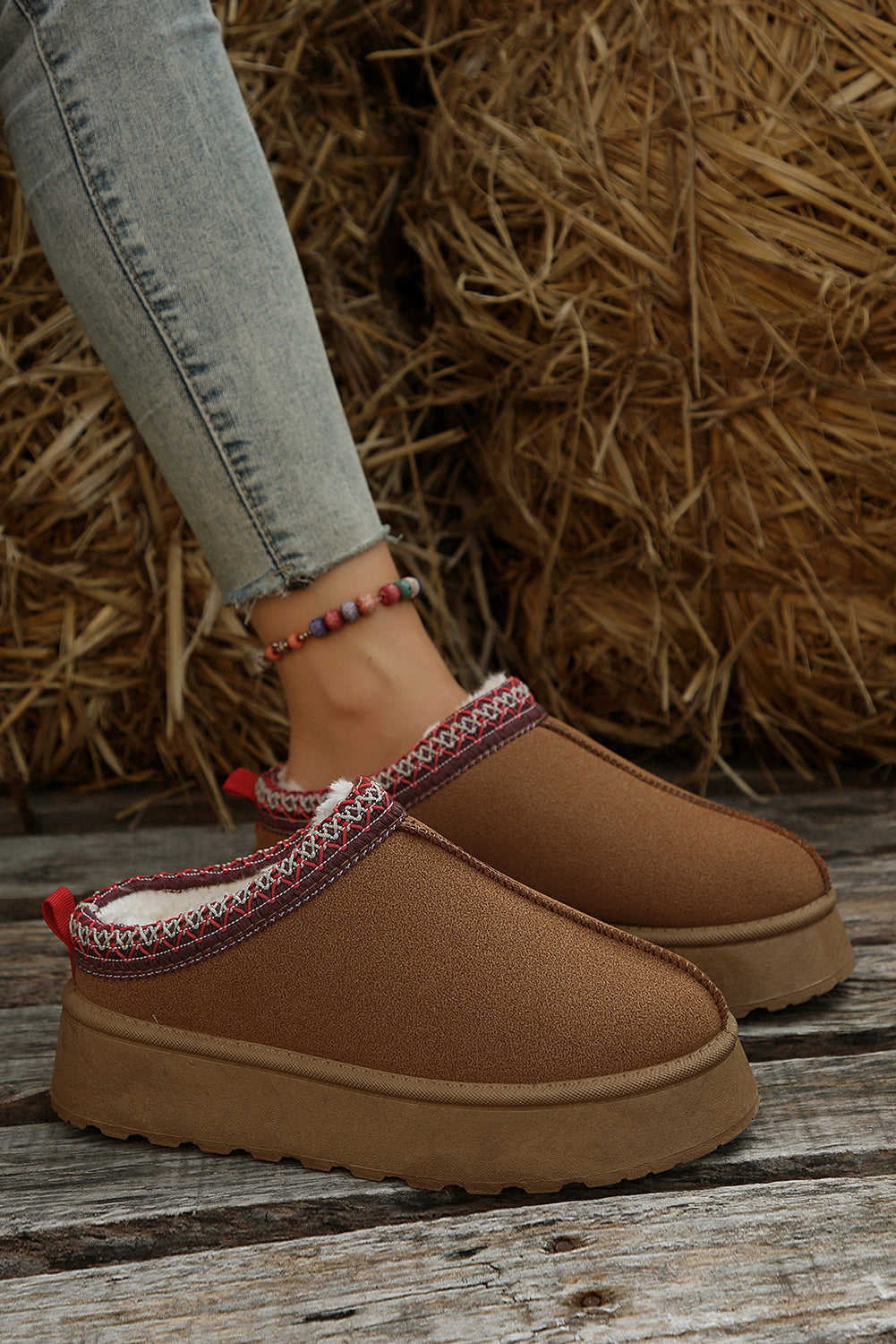 Brown slip-on shoes with decorative trim worn by a person on a wooden floor with hay in the background.