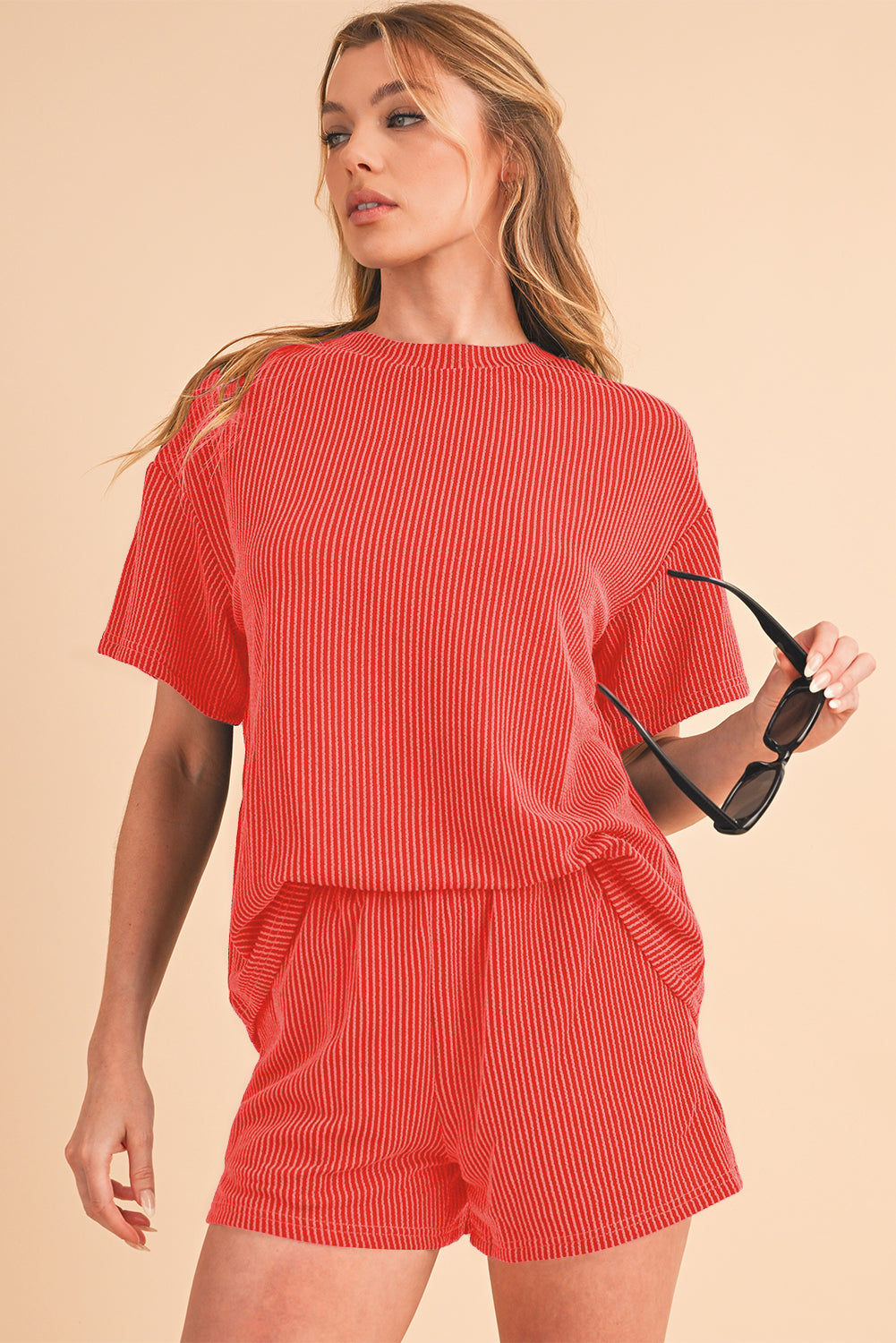 Woman wearing a red ribbed outfit holding sunglasses on a beige background