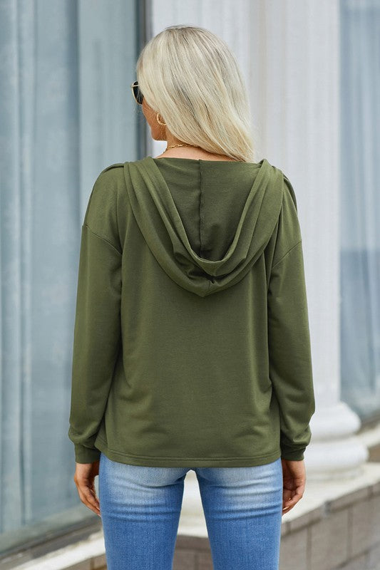 Person wearing an olive green hoodie and blue jeans standing against a light-colored wall.