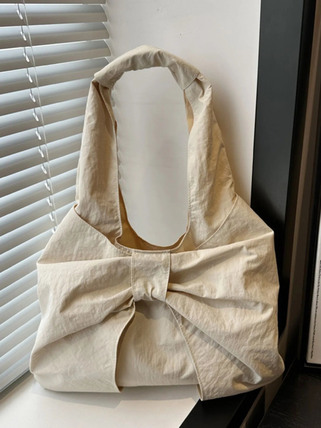 Beige handbag with a bow detail on a surface near a window with blinds.