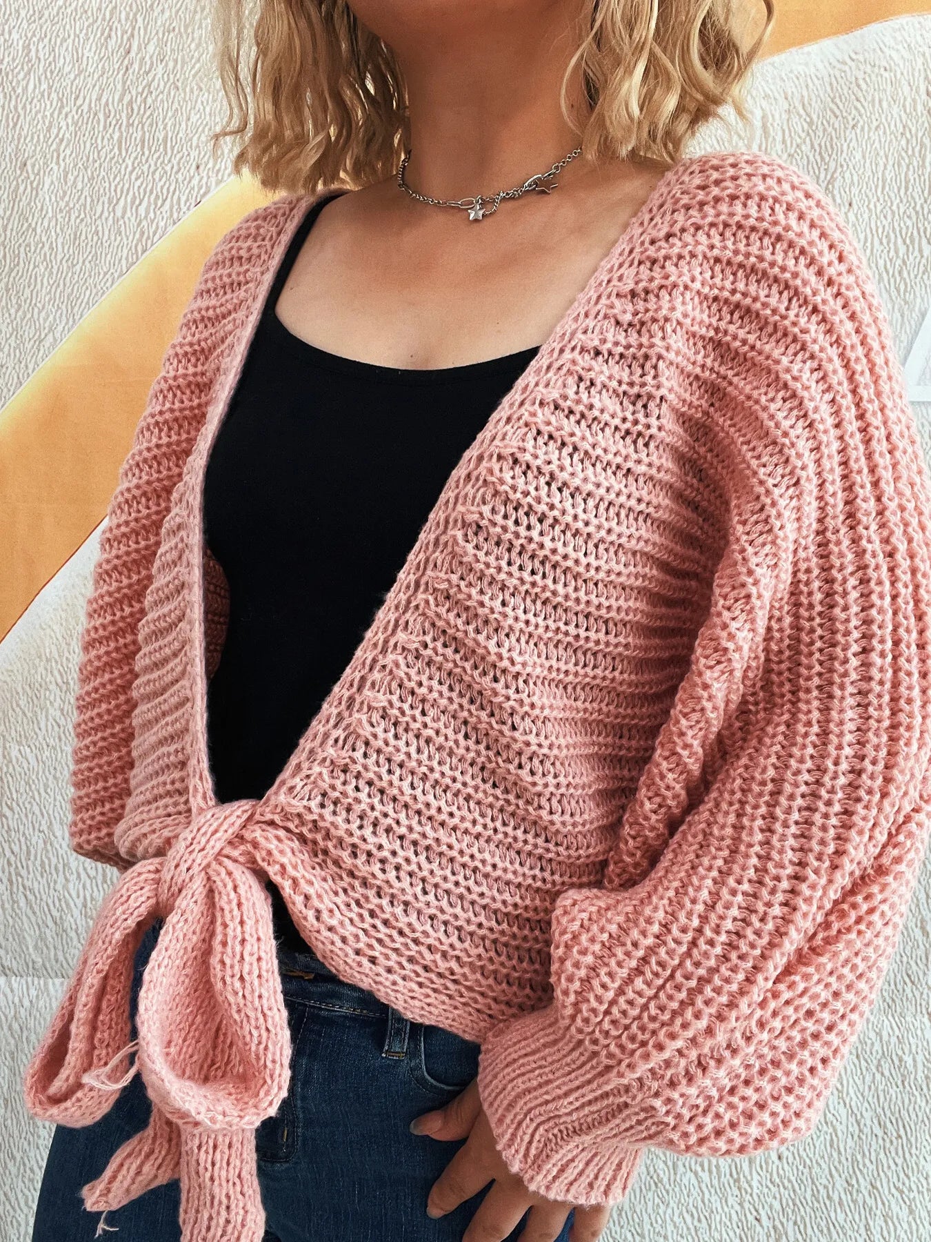 Person wearing a pink knitted cardigan over a black top and blue jeans on a neutral background